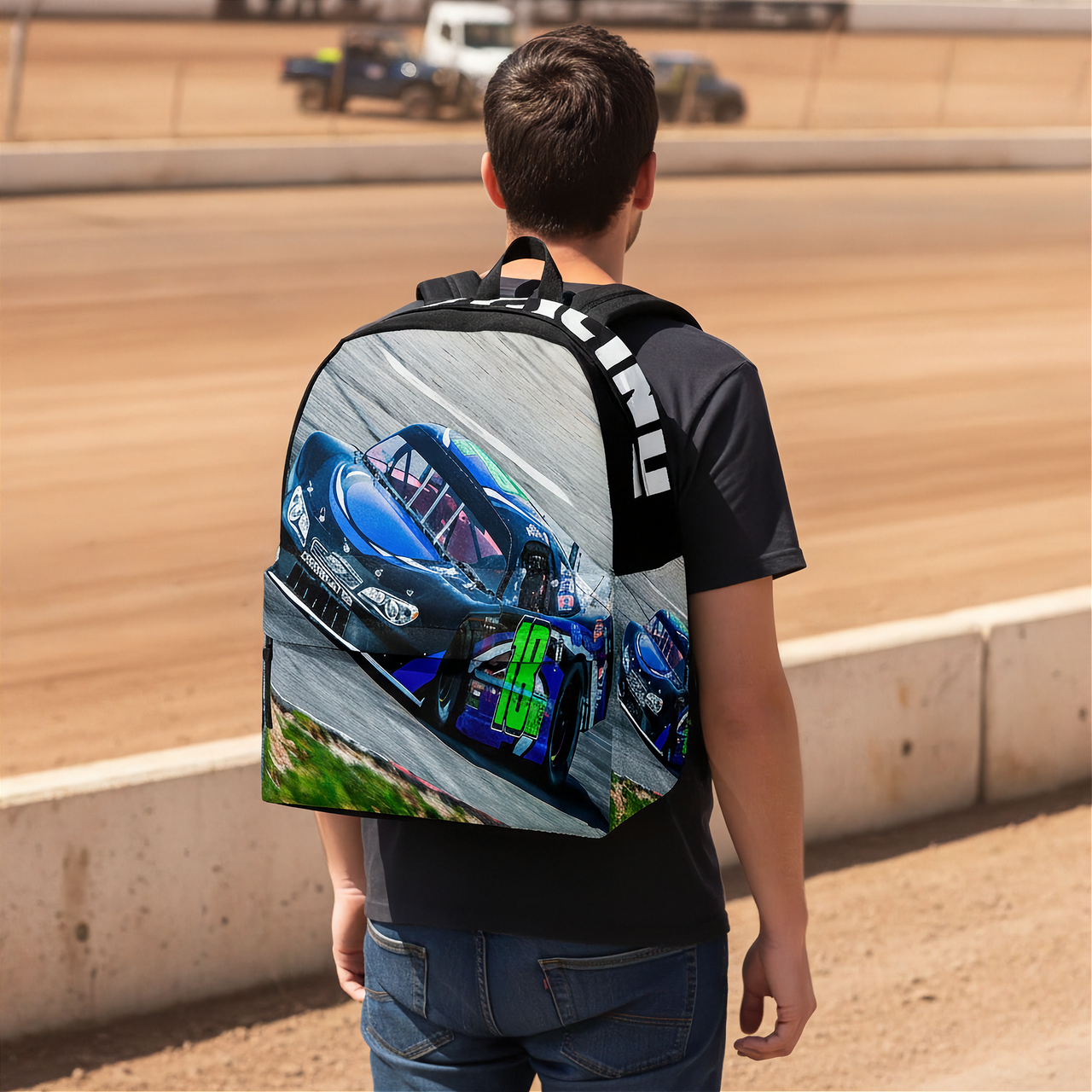 A man viewed from behind wearing a black backpack printed with a high-speed racing scene featuring blue and green late model car on a track. The background shows a blurred dirt racetrack.