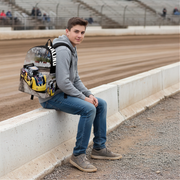 A young man sitting on a concrete barrier at a racetrack, wearing a custom printed backpack featuring yellow and blue late model race car with the word 'racing' on the side.