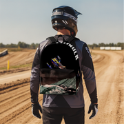 A person wearing a black and gray motocross jersey, helmet, and gloves stands on a dirt track wearing a backpack. The backpack features a black upper section with a graphic of a motocross rider and the word 'CYMILIN' in white, and a lower section showing a green and gray blurred landscape.