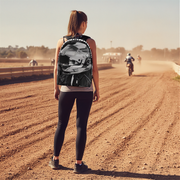 A woman stands on a dirt racetrack seen from behind, wearing a black backpack with a white and gray motorcycle graphic and the words 'Ride Safe' on the back. A motorcycle rider is visible in the dusty distance under a bright sky.