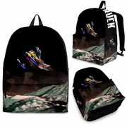 A black backpack shown from multiple angles on a white background, featuring a motocross rider on a snowmobile jumping over a snowy mountain landscape. The backpack has black straps and a white vertical text print reading 'MUCK' on the side.