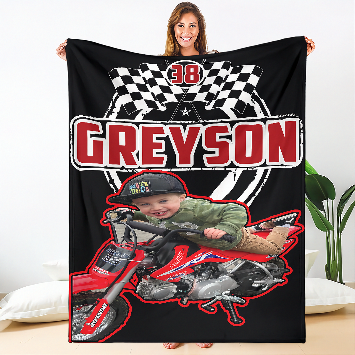 A woman holds up a black custom blanket featuring a racing design with the name 'GREYSON' in large red letters, checkered flags, a number 38, and a photo of a child riding a red dirt bike. The background is a brightly lit room with white walls and a plant.