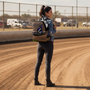 A woman stands on a dirt track wearing a backpack with non wing sprint race car. The backpack has a wood-grain patterned body with black straps and white text reading 'SPECIALTY'.