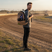 A man standing on a dirt racetrack at sunset wearing a large, custom-designed backpack. The backpack features a wood-grain pattern on the sides and a black center panel with a blue and black remote control car graphic.
