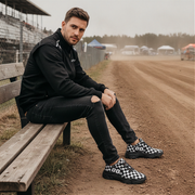 Man sitting on a wooden bench at a race track wearing black and white checkered shoes.