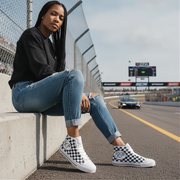 Woman sitting on a race track wearing checkered sneakers with a car in the background