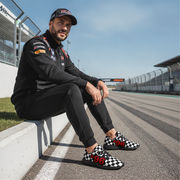 Man sitting on a race track wearing checkered shoes with numbers, black outfit, and cap.