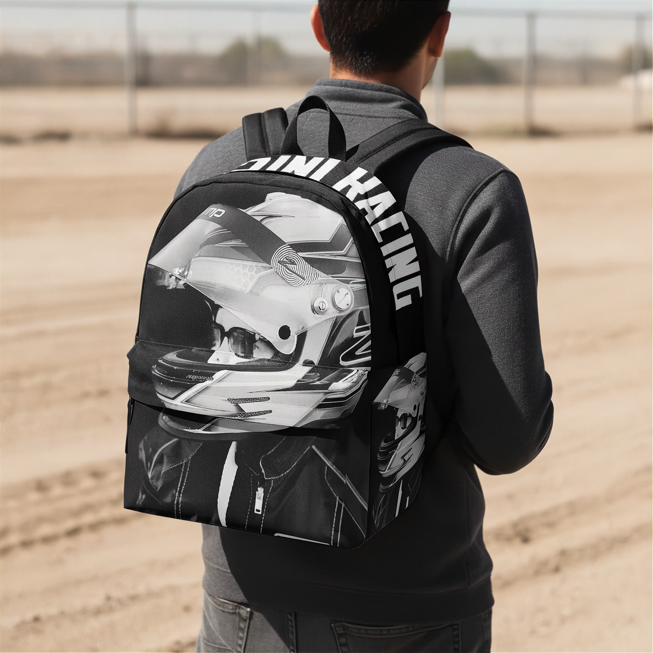 A person viewed from behind wearing a black backpack with a large black-and-white racing helmet graphic and the words 'INI RACING' across the back, standing in a sandy outdoor area.