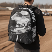 A man viewed from behind wearing a black backpack with a large grayscale racing helmet graphic and the word 'RACING' printed vertically on the back, standing on a dirt track.