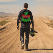 A man viewed from behind walking on a dirt road wearing a black motocross jacket with a large red and green graffiti-style graphic on the back, black pants, and holding a black and green helmet.