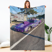 A woman holds up a large printed blanket or throw featuring a purple race car on a racetrack, with a white trailer in the background. The blanket is displayed against a clean indoor backdrop with pillows and a plant visible.