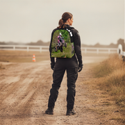 A woman stands on a dirt path outdoors, viewed from behind, wearing a black riding jacket and pants with a colorful graphic print on the back showing a dirt bike rider in a grassy field.