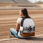 A woman sits on the edge of a dirt track wearing a backpack with a printed graphic of a sprint car, set against a blurred background of stadium seating.