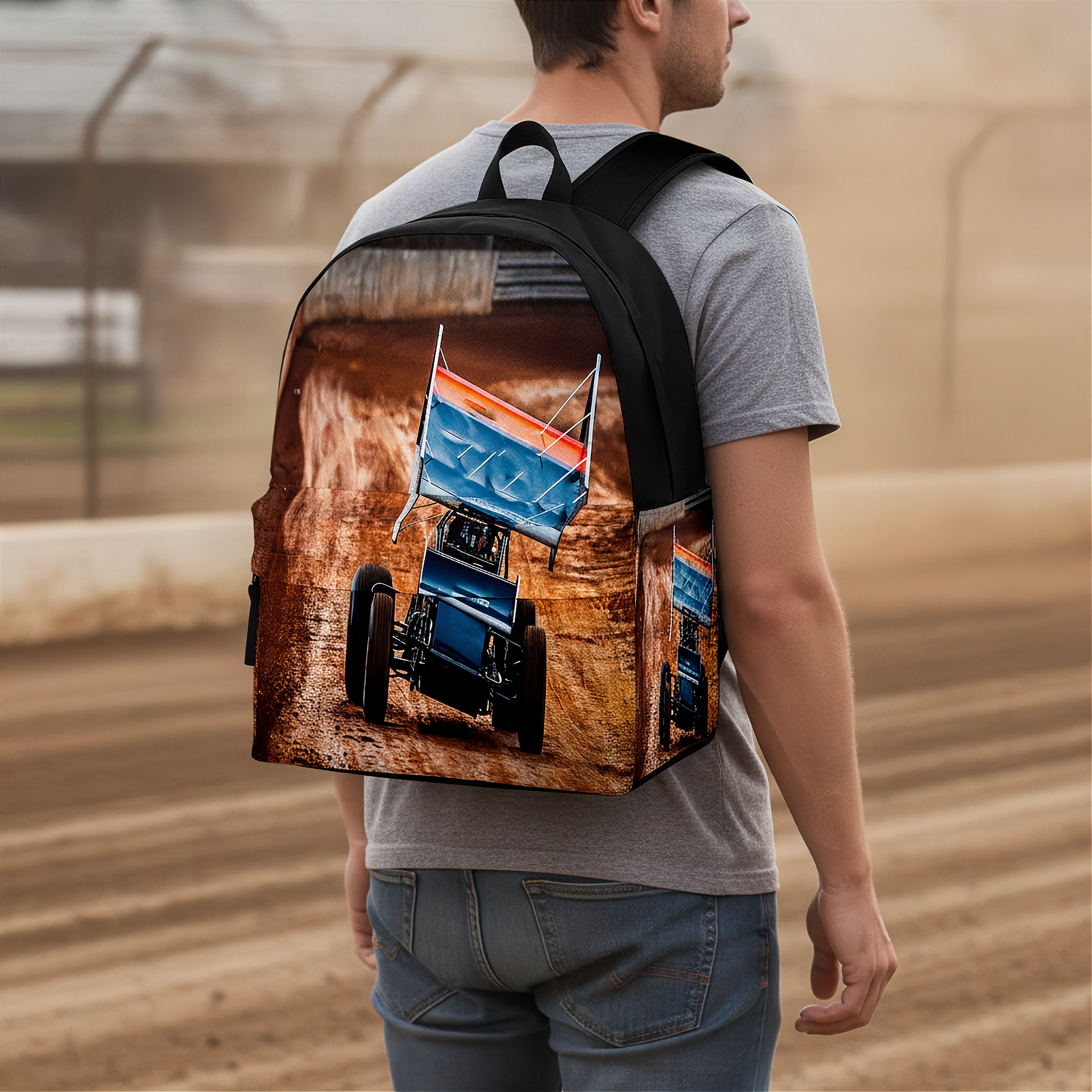 A man wearing a gray t-shirt and blue jeans is shown from behind, carrying a backpack with a printed graphic of a blue sprint race car on a dirt track with a flag that reads 'GO!'. The background is a blurred dirt racetrack.