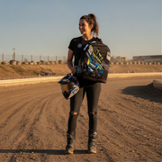 A woman stands on a dirt racetrack at golden hour wearing a black t-shirt and ripped black jeans, holding a motorcycle helmet in one hand and wearing a large black backpack with a colorful blue, yellow, and white racing graphic in the other hand.