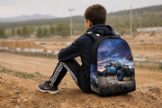 A boy wearing a black hoodie and black track pants sits on a dirt hill, looking out over a dirt track. He is wearing a black backpack with a large graphic print of a blue and black off-road vehicle on a dramatic sky background.