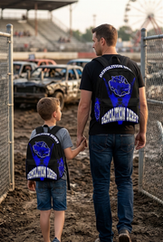 A young boy aged 10 wearing a custom blue demolition derby backpack standing near a muddy fairground arena fence, watching smashed derby cars in the distance with an adult woman by his side.