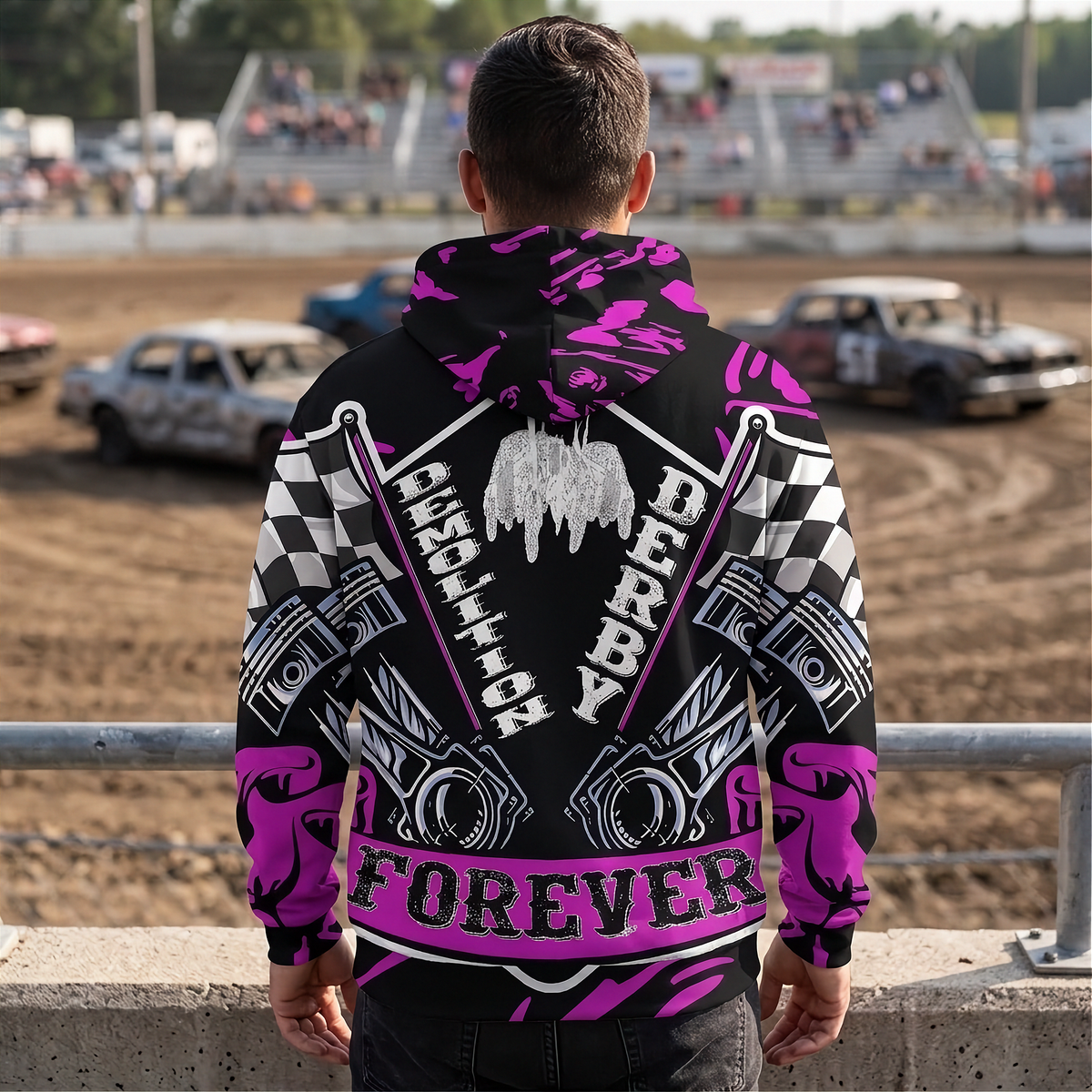 A man stands with his back to the camera wearing a black hoodie with a highly detailed, colorful racing graphic print featuring pink, white, and black. The background shows a dirt track with race cars and spectators in the stands.
