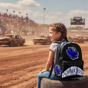 A young girl sits on a tire at a dirt racing track, wearing a black backpack with blue and white graphics that read 'Demolition Derby' and show a blue ram head logo. Dirt cars race in the background under a cloudy sky.