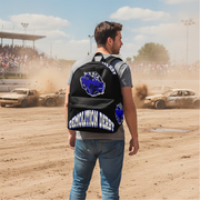 A man stands on a dirt race track wearing a black backpack with a blue and white 'Demolition Derby' graphic, with race cars and a crowd in the background.