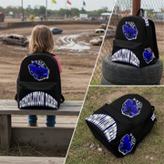 A black backpack with a blue and white 'Demolition Derby' graphic, shown in a three-panel collage. One panel shows a child sitting on a wooden bench looking out toward a dirt track, while the other two panels show the backpack from different angles, including on tires and on the grass.