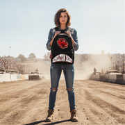 A woman stands on a dusty racetrack wearing a black denim jacket with a red and white 'demolition derby' graphic on the front, paired with ripped blue jeans and brown boots.