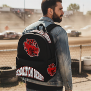 A man wearing a denim jacket stands with his back to the camera, showcasing a black backpack with red and white 'Demolition Derby' graphics and patches in an outdoor dirt track setting.