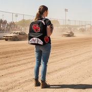 A woman stands on a dirt racetrack wearing a black embroidered biker-style vest with red and white graphics, blue jeans, and brown boots, with race cars and a crowd visible in the background.