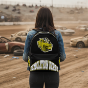 A person viewed from behind wearing a black backpack with a large yellow wrecked-car graphic and the words 'DEMOLITION DERBY' on the back, standing in a dusty racetrack setting with cars and bleachers in the background.