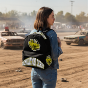 A woman stands in a dusty desert racetrack wearing a black backpack with 'Demolition Derby' graphics and patches on the back, alongside a distressed car in the background.