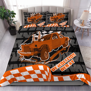 A bed dressed in a printed bedding set featuring an orange race car, black-and-white checkered stripes, and the words 'Demolition Derby' in a bold racing-style font. The set includes a duvet cover and two matching pillowcases, shown in a styled bedroom scene with a headboard, side table, lamp, plant, and window.