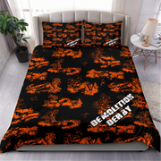 A made bed in a bedroom setting featuring a black bedding set with an orange graffiti-style pattern of cars and the words 'DEMOLITION DERBY' in white. The room includes a gray upholstered headboard, a side table with a lamp, a potted plant, and light wood flooring.