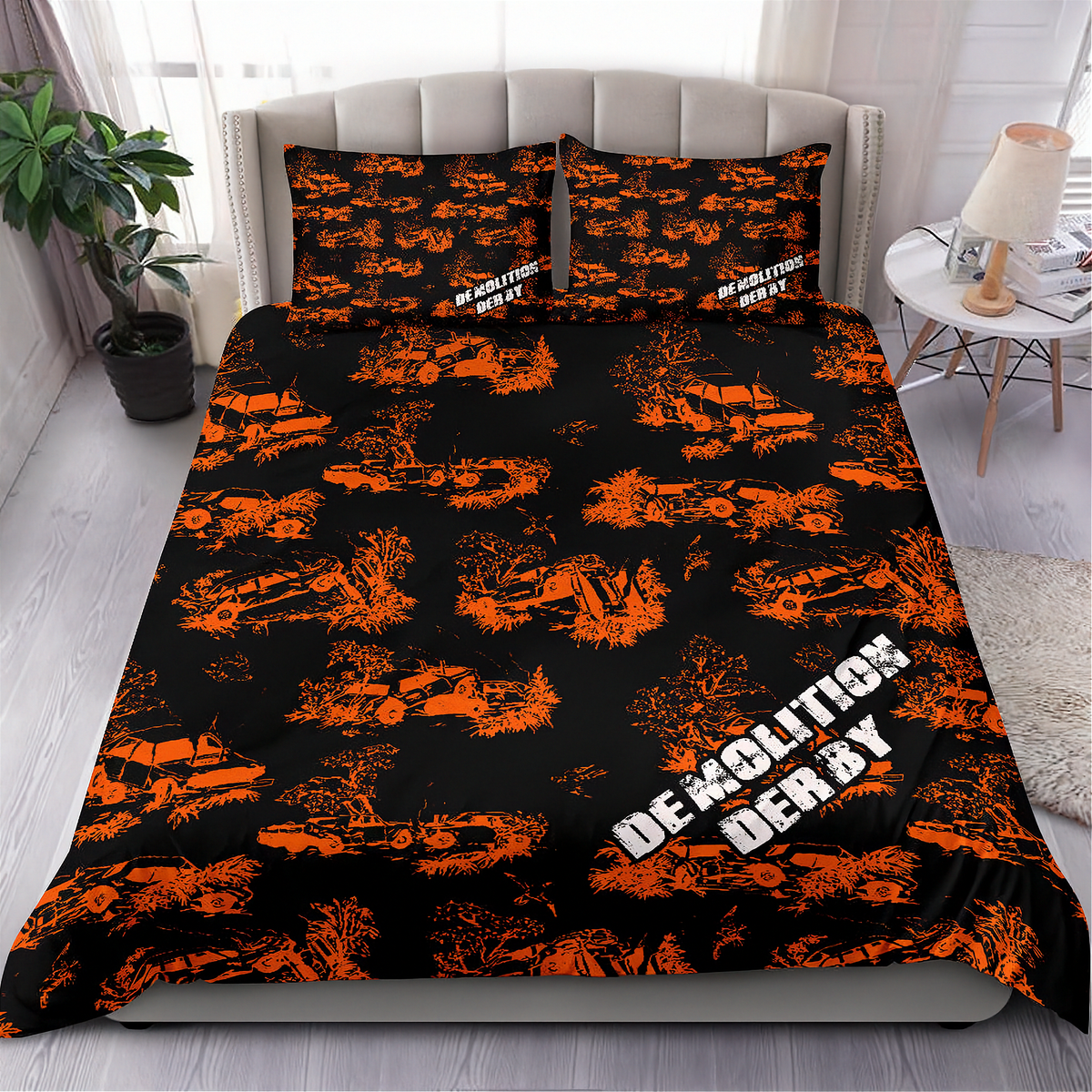 A made bed in a bedroom setting featuring a black bedding set with an orange graffiti-style pattern of cars and the words 'DEMOLITION DERBY' in white. The room includes a gray upholstered headboard, a side table with a lamp, a potted plant, and light wood flooring.