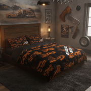 A bed dressed in a black and orange distressed racing-themed bedding set, styled in a rustic, garage-washed bedroom with racing posters, a wall lamp, and a tire decoration.
