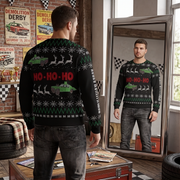 A man stands in a garage-like room wearing a black Christmas sweater with green and red holiday graphics, shown from the back while a mirror reflects his front view. The sweater features 'HO-HO-HO' text, reindeer, snowflakes, and green truck illustrations.