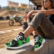A woman sits on a concrete barrier at a dirt track wearing a pair of low-top sneakers with a black and bright green graphic design featuring a motorcycle motif. A green and white dirt bike is blurred in the background.