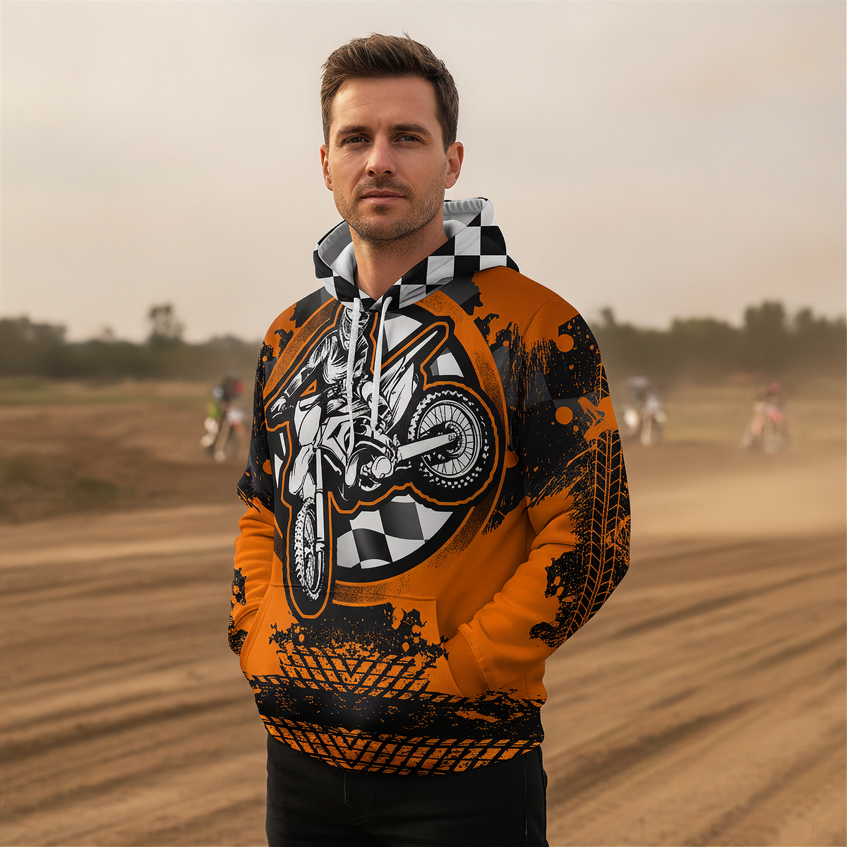 A man wearing a vibrant orange and black graphic hoodie with a motorcycle design, standing in a dusty dirt track environment.
