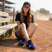 Woman sitting on a dirt track wearing black checkered shoes with blue accents and a black cap.