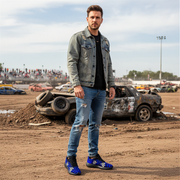 Man standing in a demolition derby area with damaged cars in the background wearing a blue muddy checkered sneakers