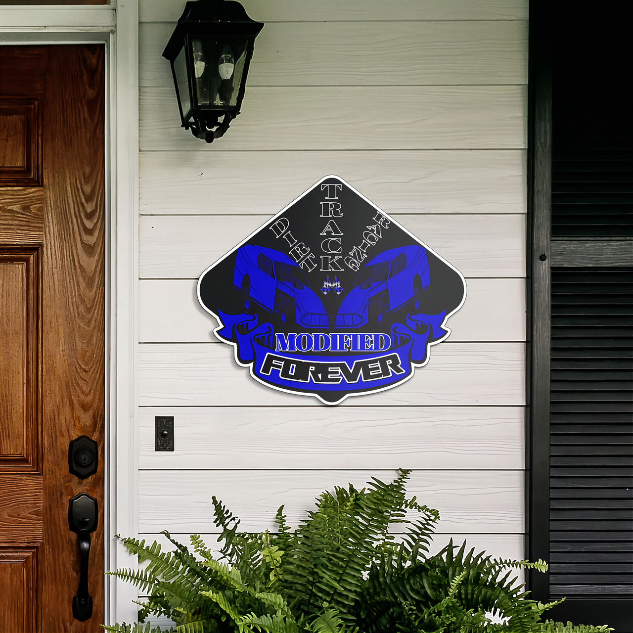 A custom diamond-shaped metal sign mounted on a white house exterior wall. The sign features a black background with a blue and silver stylized muscle car graphic and the text 'TRACK BACK' and 'MODIFIED FOREVER'.