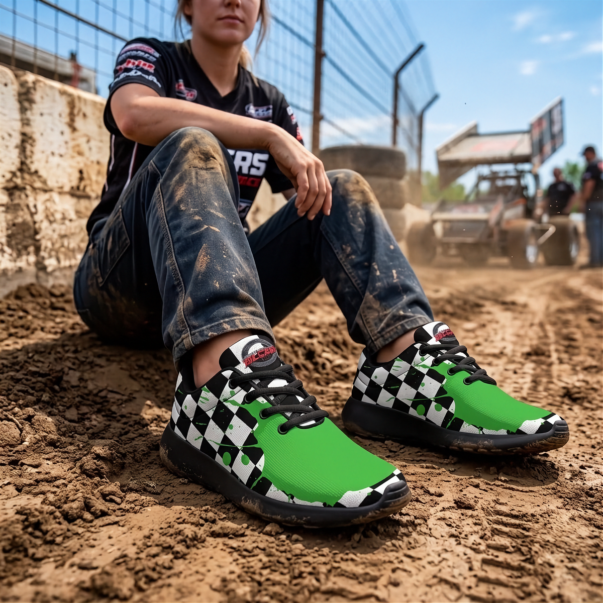 A woman sitting on dirt at an outdoor track wearing green, black, and white checkered sneakers, with a blurred racetrack and vehicle in the background.