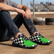 A person sitting on a dirt track wearing a pair of athletic sneakers with a black and white checkered pattern on the sides and bright green uppers, paired with dark blue jeans.