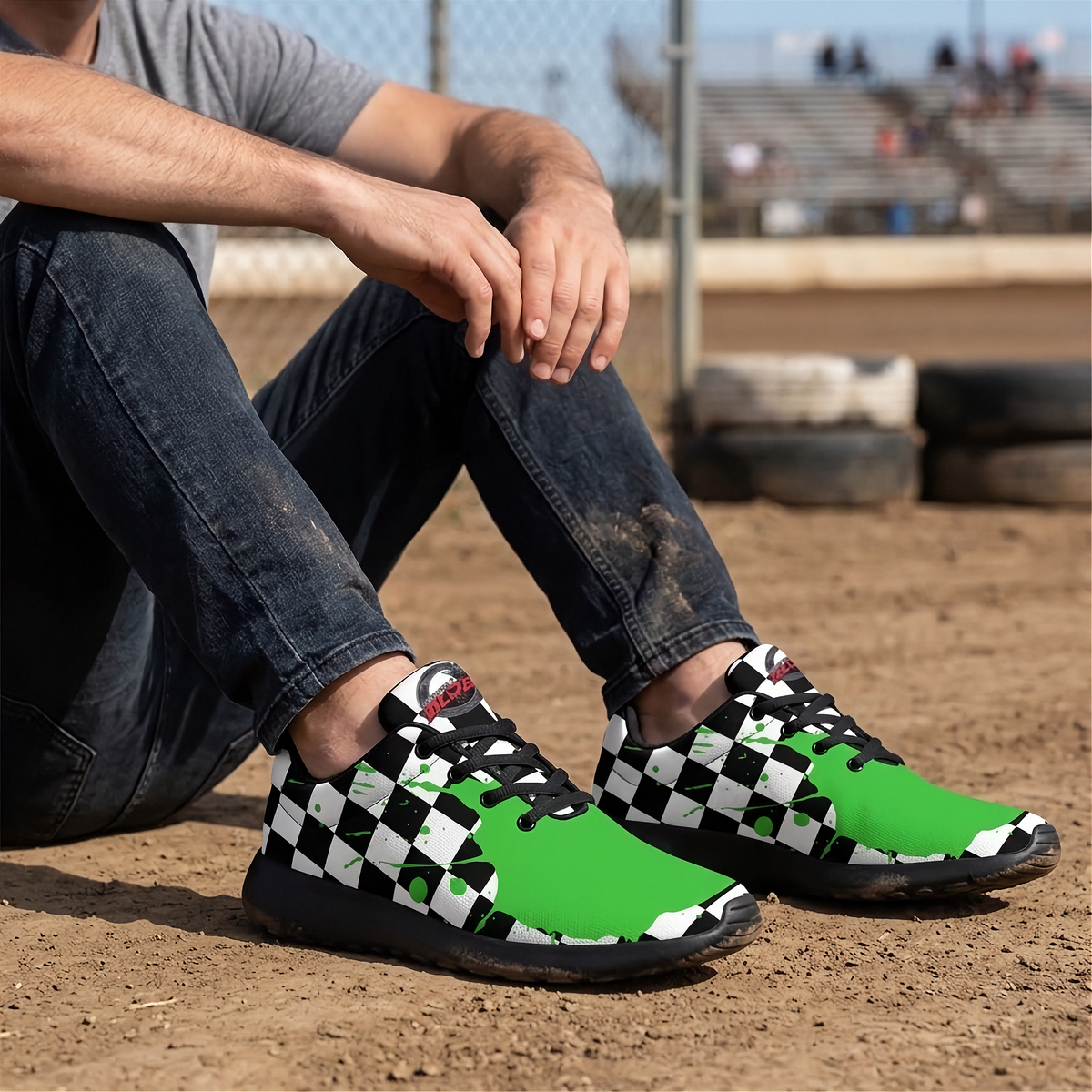 A person sitting on a dirt track wearing a pair of athletic sneakers with a black and white checkered pattern on the sides and bright green uppers, paired with dark blue jeans.