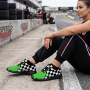 A woman sits on a racetrack wearing black checkered and bright green sneakers with black socks and black athletic pants.