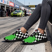A person sitting on a racetrack barrier wearing green and black checkered sneakers with a green checkered flag graphic on the side. The background shows a blurred racetrack scene with a lime green race car and pit stop signage.