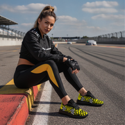 Woman in black and yellow racing outfit with yellow checkered sneakers sitting on a track..