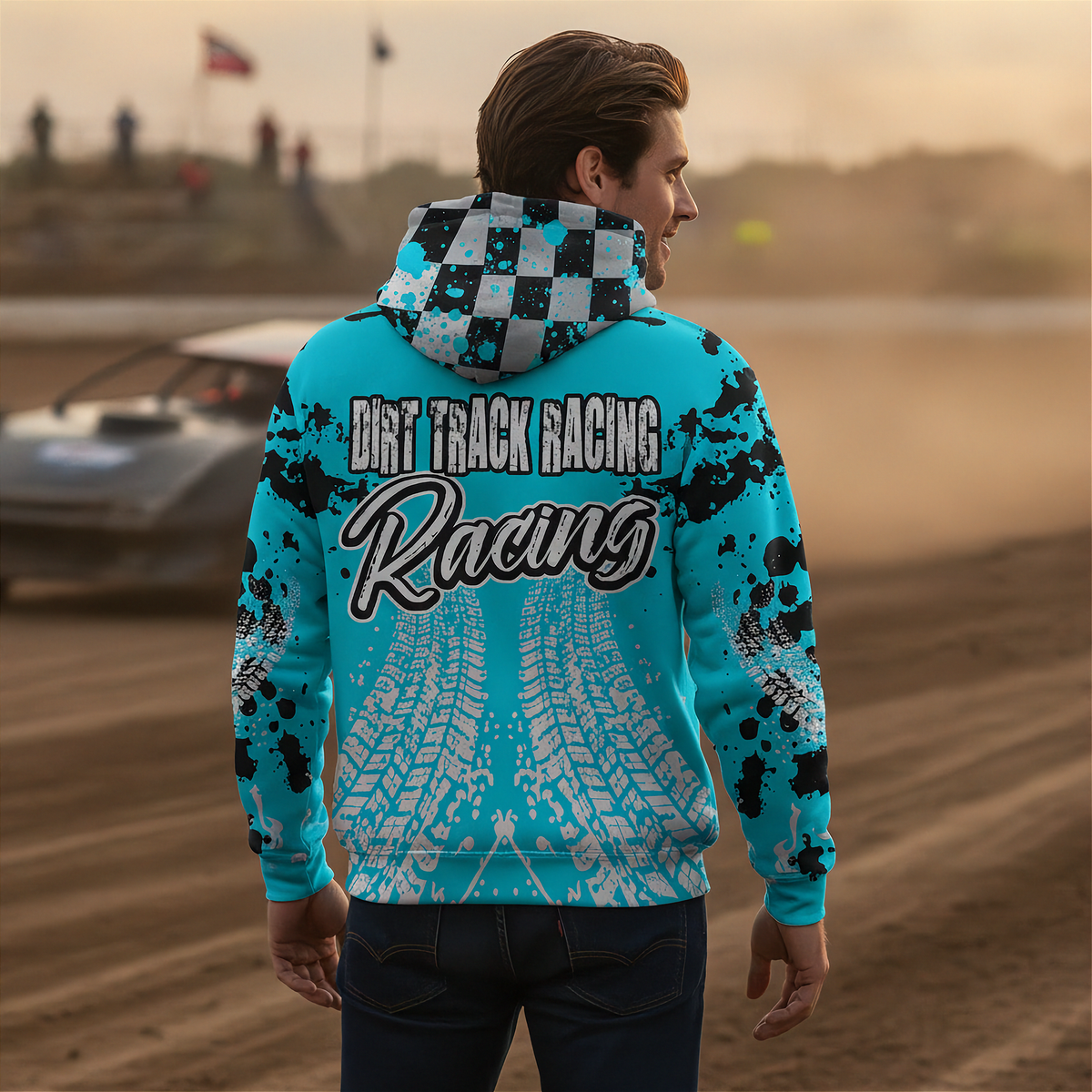 A man stands on a dirt track wearing a bright turquoise hoodie with black splatter graphics, a black-and-white checkered hood, and large 'Dirt Track Racing' text across the back, with a blurred race car and track in the background.