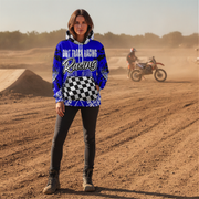 A woman stands on a dirt track wearing a blue and black checkered hoodie with 'Dirt Track Racing' printed on the front, paired with black pants and boots. A dirt bike rider is visible in the dusty background.