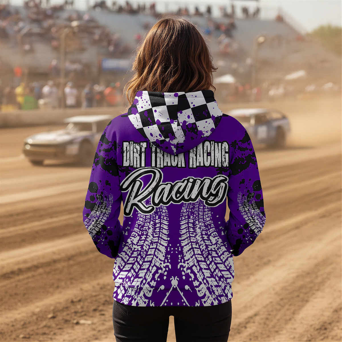 A person standing on a dirt track wearing a purple and black hooded sweatshirt with 'Dirt Track Racing' and tire tracks printed on the back, with race cars and a blurred trackside crowd in the background.