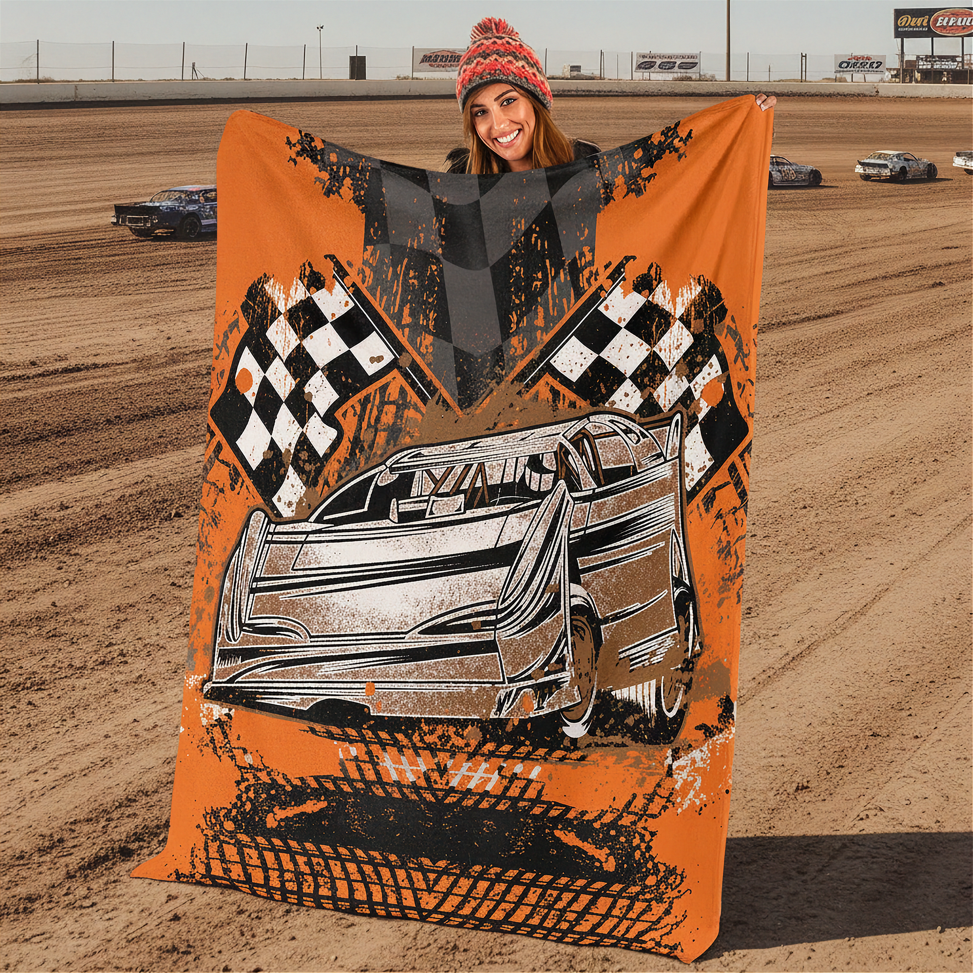 Woman holding a racing-themed blanket with a car and checkered flags design at a dirt track.
